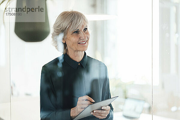 Lächelnde Geschäftsfrau mit Tablet-PC hinter Glasscheibe im Büro
