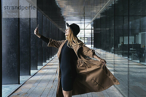 Frau hält Trenchcoat in der Hand und macht ein Selfie mit dem Smartphone auf dem Fußweg