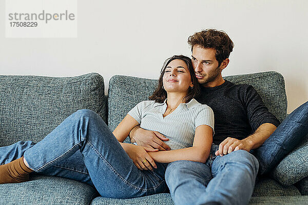 Lächelndes junges Paar  das beim gemeinsamen Sitzen auf dem Sofa nachdenkt