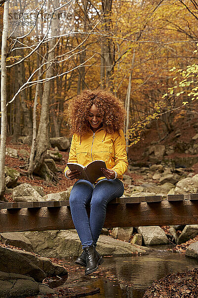 Lächelnde junge Frau  die ein Buch liest  sitzt auf einer Brücke im Herbstwald