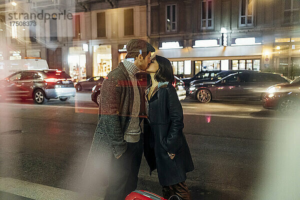 Junges Paar in warmer Kleidung küsst sich auf beleuchteter Stadtstraße