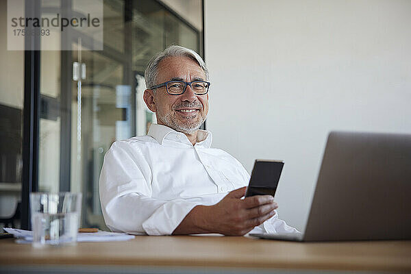Lächelnder reifer Geschäftsmann mit Smartphone und Laptop im Büro