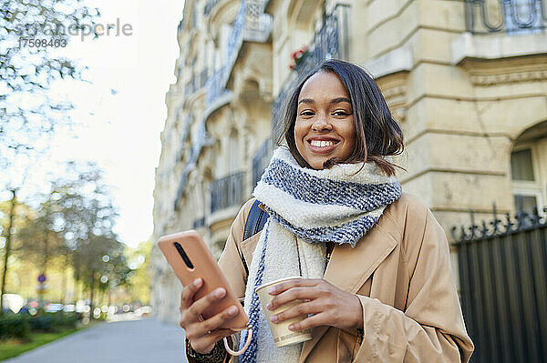 Lächelnde Frau mit Mobiltelefon und Einwegbecher in der Stadt