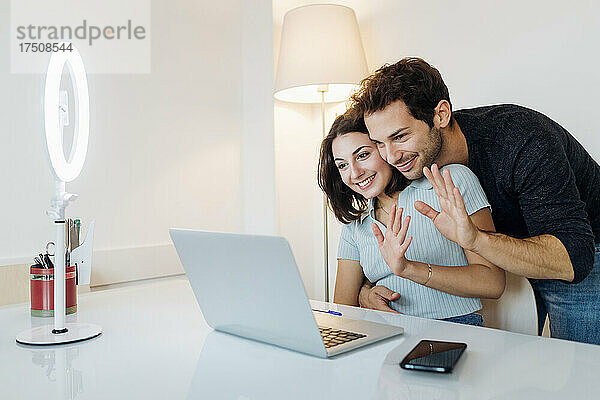 Lächelndes junges Paar winkt zu Hause per Videoanruf über den Laptop