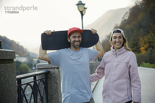Mann hält Skateboard und Frau steht neben Geländer auf Fußweg