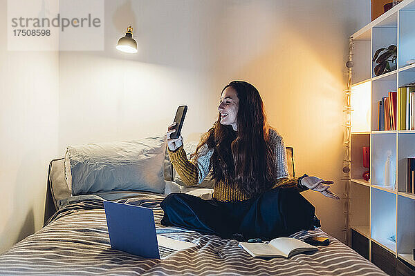 Frau macht Videoanruf über Smartphone und sitzt mit Laptop zu Hause im Bett