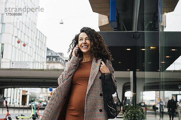 Lächelnde Geschäftsfrau  die beim Spaziergang in der Stadt mit ihrem Smartphone telefoniert