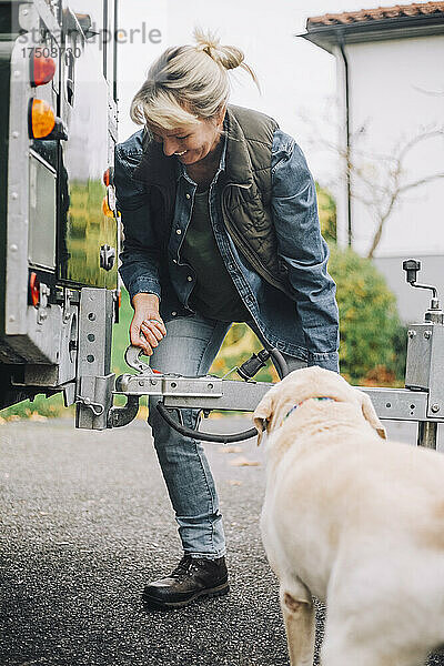 Lächelnde Frau  die einen Anhänger an einem Geländewagen mit Hund in der Einfahrt zieht