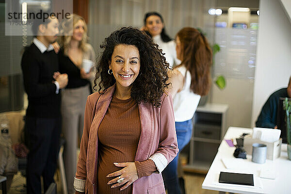 Porträt einer lächelnden schwangeren Geschäftsfrau mit Kollegen im Hintergrund im Büro