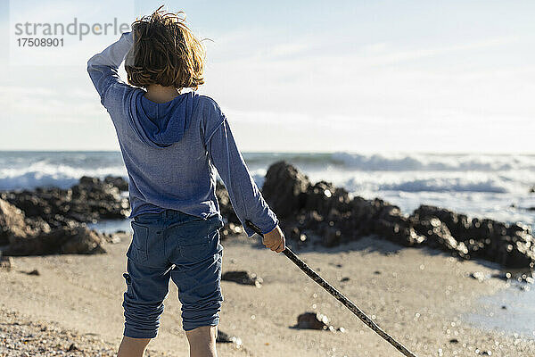 Junge spielt an einem felsigen Strand und hält einen langen Seetangstrang in der Hand
