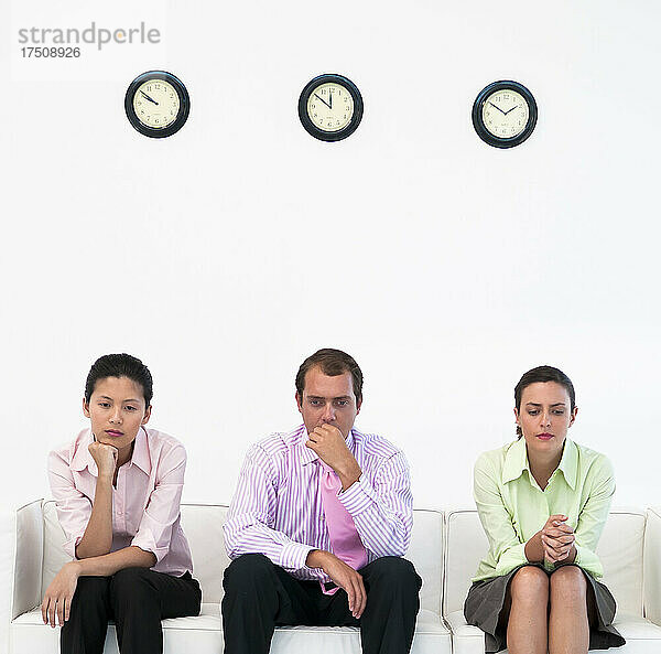 Drei Geschäftsleute sitzen auf einer Couch unter einer Uhr und wirken nervös.