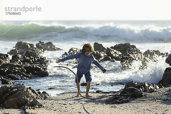 Kleiner Junge spielt im Sand zwischen Felsen am Strand  die Wellen brechen