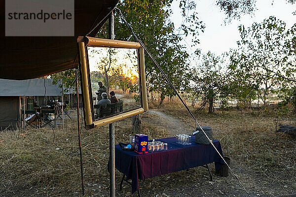 Ein Safarilager  Zelte und Getränketisch  ein großer Spiegel an einer Zeltstange  drei Personen bei Sonnenuntergang