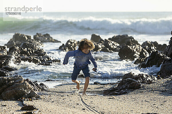 Kleiner Junge spielt im Sand zwischen Felsen am Strand  die Wellen brechen