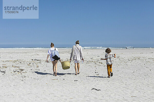 Mutter und Tochter im Teenageralter gehen an einem Sandstrand spazieren und tragen einen Korb  ein Junge folgt.