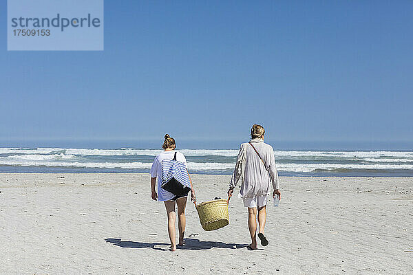 Mutter und Tochter im Teenageralter gehen am Sandstrand spazieren und tragen einen Korb
