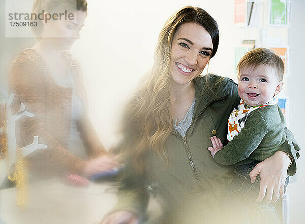 Frau mit Baby neben einer Registrierkasse mit Verkäuferin  die in die Kamera lächelt.