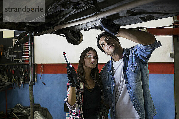 Junge Frau und Mann arbeiten im Team an einer Autowerkstatt  stehen unter dem Auto am Nivelliersystem und diskutieren über Reparaturen