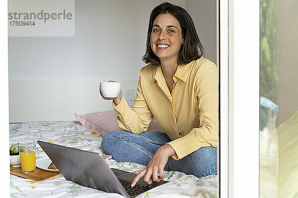 Lächelnde Frau trinkt Kaffee  während sie im Schlafzimmer einen Laptop benutzt