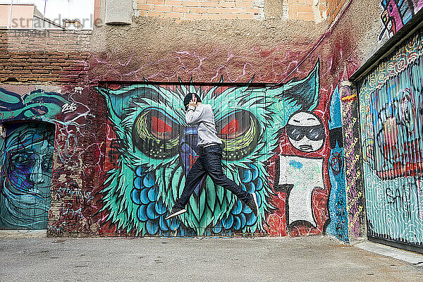 Junger Mann mit Kopfhörern springt vor Graffiti in die Luft