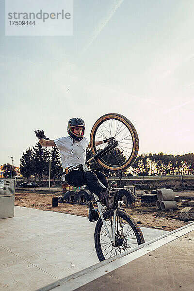 Sorgloser Mann mit Helm führt bei Sonnenuntergang einen Stunt mit dem Fahrrad auf der Rampe im Park durch