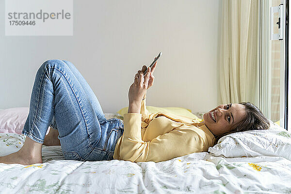 Lächelnde Frau benutzt Mobiltelefon  während sie im Schlafzimmer liegt