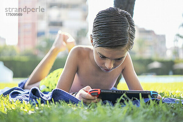 Hemdloser Junge spielt Videospiel über digitalem Tablet  während er auf einem Handtuch im Garten liegt