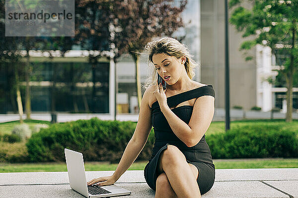Geschäftsfrau spricht mit Smartphone  während sie im Freien einen Laptop benutzt