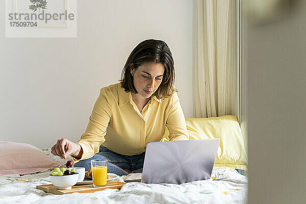 Frau frühstückt und benutzt Laptop im Schlafzimmer