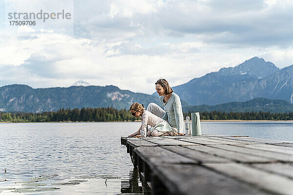 Mutter mit Tochter sitzt auf dem Steg über dem See vor dem Himmel