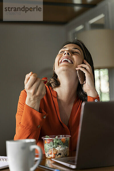 Glückliche Geschäftsfrau telefoniert  während sie zu Hause Salat isst