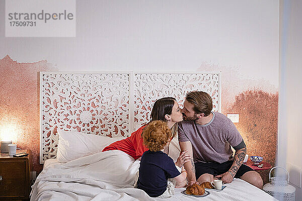 Eltern küssen sich beim Frühstück mit ihrem Sohn im Bett zu Hause