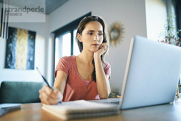 Ernsthafte Freiberuflerin schaut auf den Laptop  während sie in Notizblock auf dem Schreibtisch schreibt