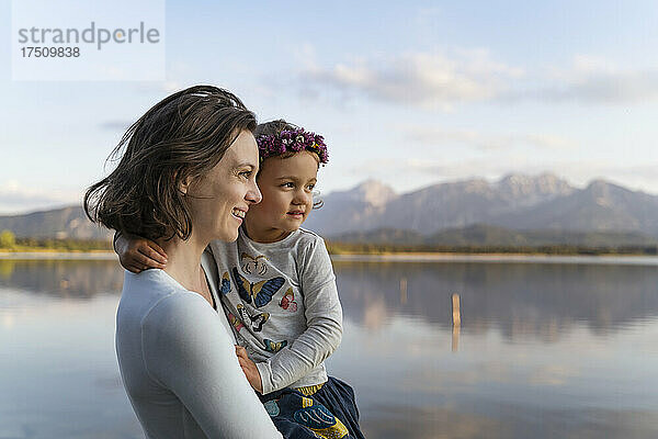 Lächelnde Mutter trägt ihre süße Tochter  während sie bei Sonnenuntergang am See steht
