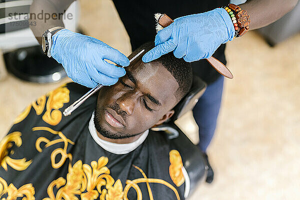 Nahaufnahme eines Friseurs mit Handschuhen  der dem Kunden im Salon mit einer Rasierklinge die Haare schneidet