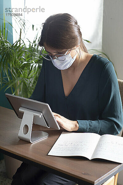 Junge Frau trägt Schutzmaske und benutzt Tablet zu Hause auf dem Schreibtisch