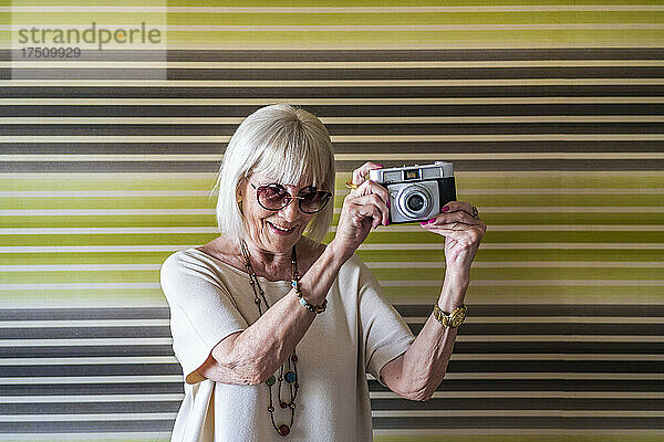 Stilvolle Seniorin mit Sonnenbrille fotografiert zu Hause mit der Kamera vor der Wand
