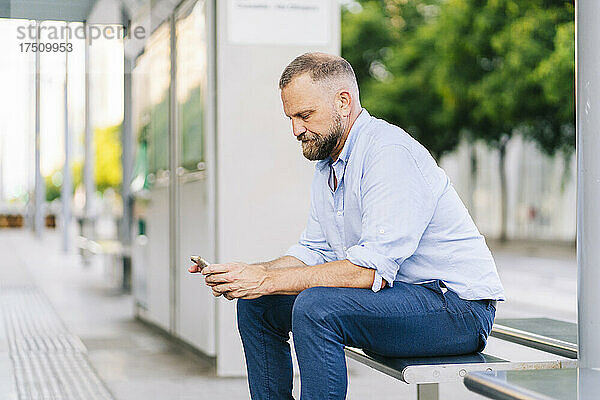 Geschäftsmann benutzt Smartphone  während er auf einer Bank am Bahnhof sitzt