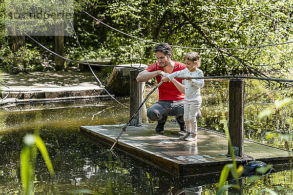 Mädchen spielt mit Stock  während sie mit ihrem Vater auf einem Holzfloß über einem See im Wald steht