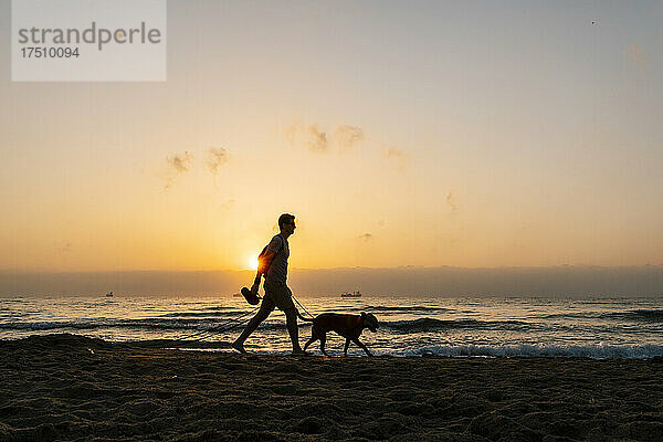 Silhouette eines Mannes  der im Morgengrauen mit seinem Hund am Strand spaziert