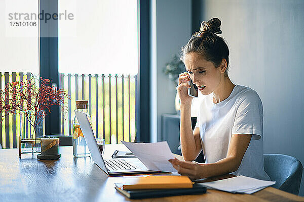 Geschäftsfrau hält Dokument in der Hand und spricht über Smartphone am Schreibtisch im Heimbüro