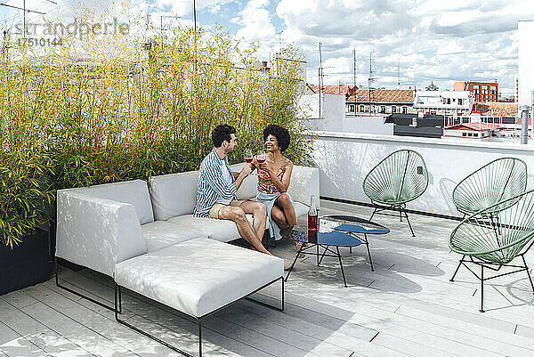 Multiethnisches Paar prostet Rotweingläser an  während es auf dem Sofa auf der Penthouse-Terrasse sitzt