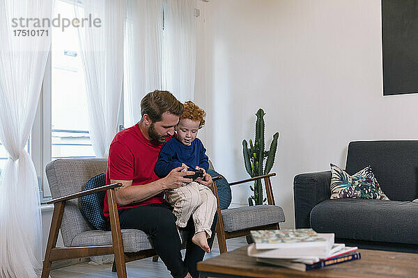 Vater und süßer Sohn halten Joystick in der Hand  während sie im heimischen Wohnzimmer Videospiel spielen