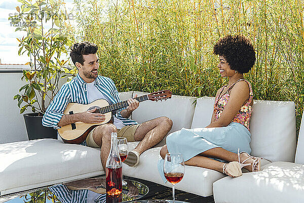 Glücklicher Mann  der für eine Frau Gitarre spielt  während er auf dem Sofa auf der Penthouse-Terrasse sitzt