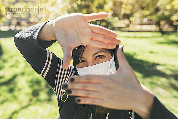 Nahaufnahme einer jungen Frau mit Maske  die im Park durch Fingerrahmen schaut