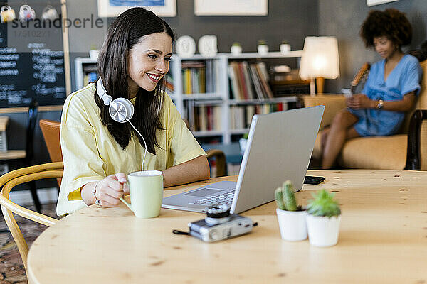 Glückliche Frau benutzt Laptop im Café mit Freundin im Hintergrund