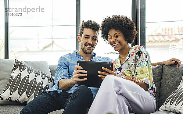 Glückliches multiethnisches Paar nutzt digitales Tablet  während es auf dem Sofa im Penthouse sitzt
