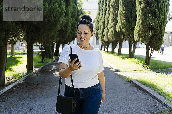 Lächelnde  kurvige junge Frau  die in einem öffentlichen Park ihr Mobiltelefon benutzt