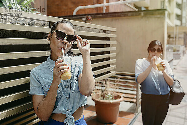 Freundinnen mit Sonnenbrille trinken Saft  während sie in der Stadt stehen