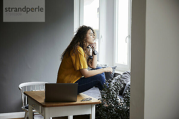 Nachdenkliche Frau blickt durch das Fenster  während sie im Wohnzimmer im Heimbüro eine Arbeitspause macht